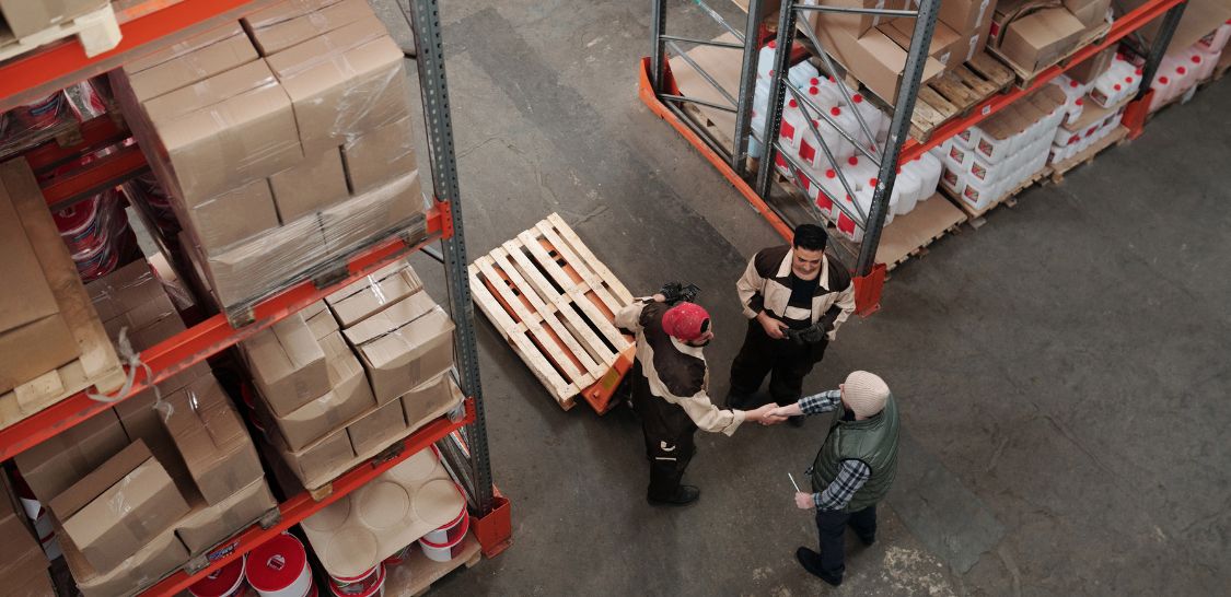 Workers standing in a warehouse with tall shelves around them are photographed from above