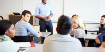 A teacher stand in front of a classroom listening to a student talking
