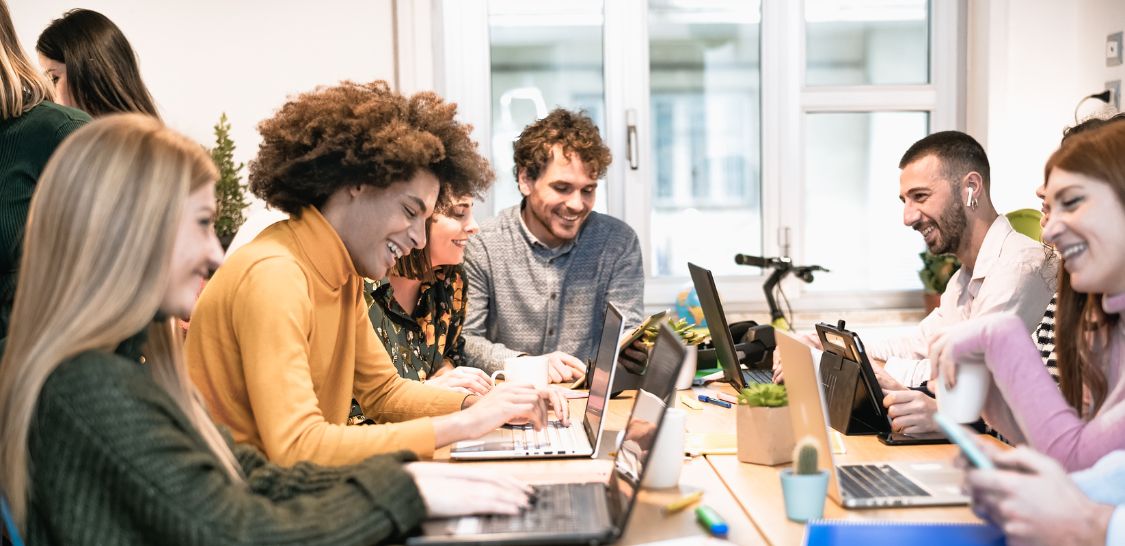 Multiple people sit at a table smiling while they work on their laptops