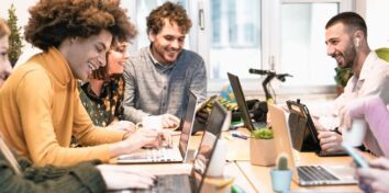 Four people sit at a table smiling while they work on their laptops