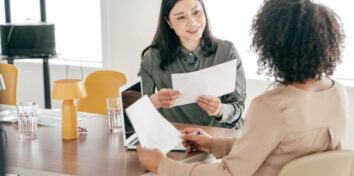 two women sit at a table across from each other, each holding a sheet of paper and there is a laptop on the table between them