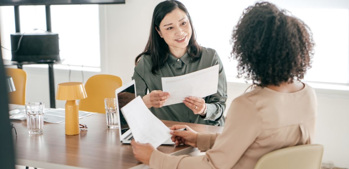 two women sit at a table across from each other, each holding a sheet of paper and there is a laptop on the table between them