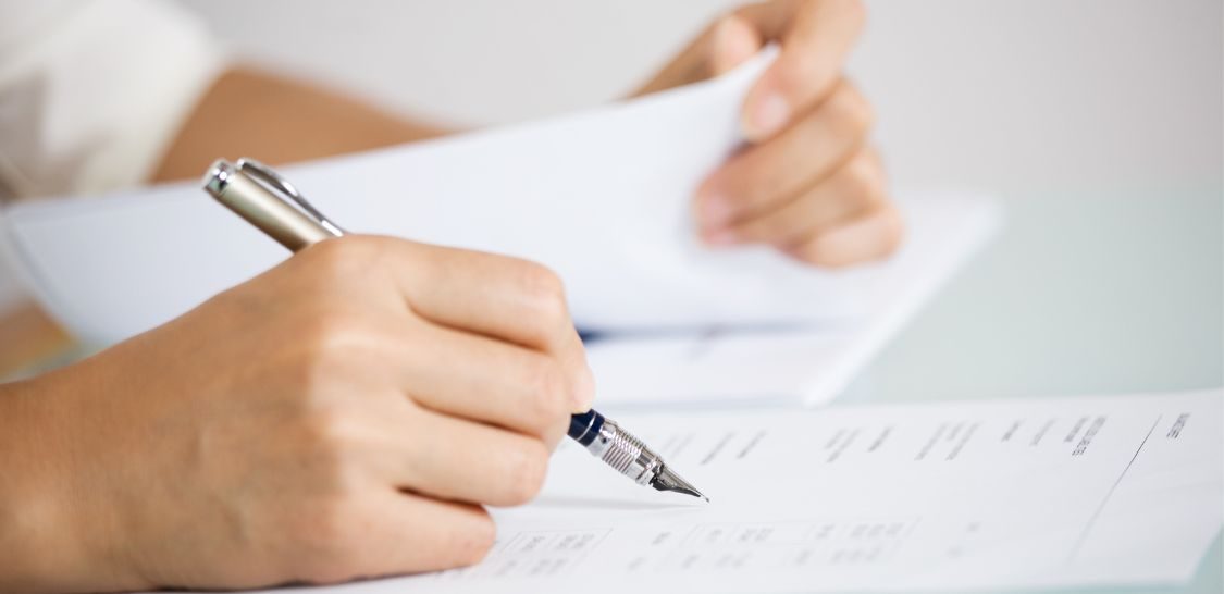 A hand holds a pen over a piece of paper with text on it while the other hand holds paper in the background