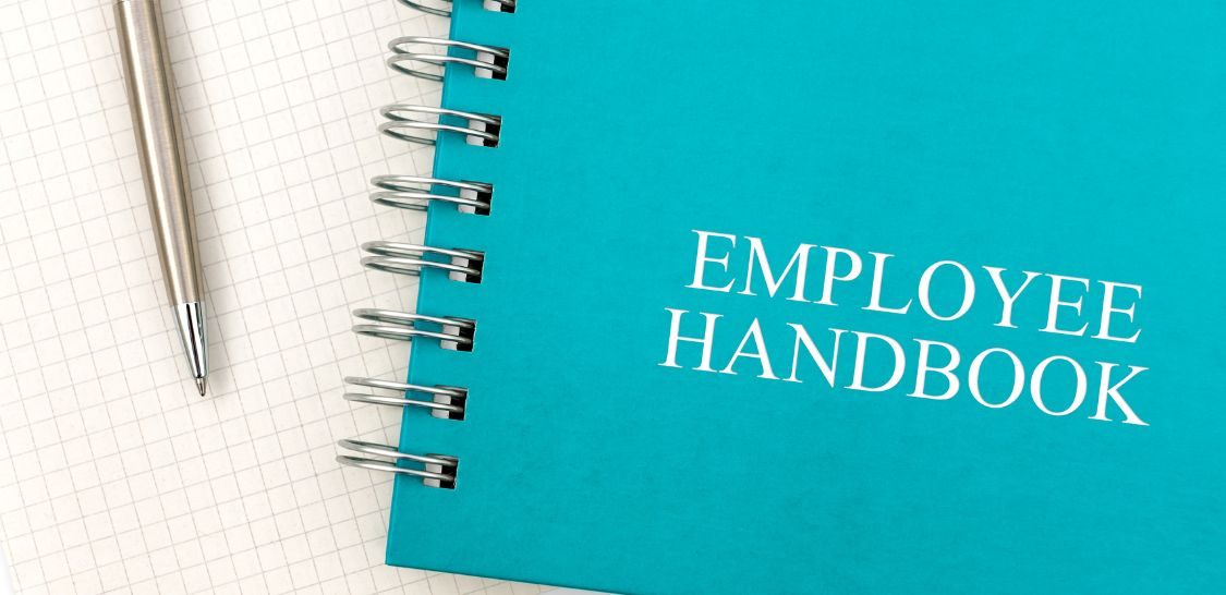 A teal spiral-bound notebook with the words "Employee Handbook" sits on a piece of graph paper with a silver pen sitting to its left