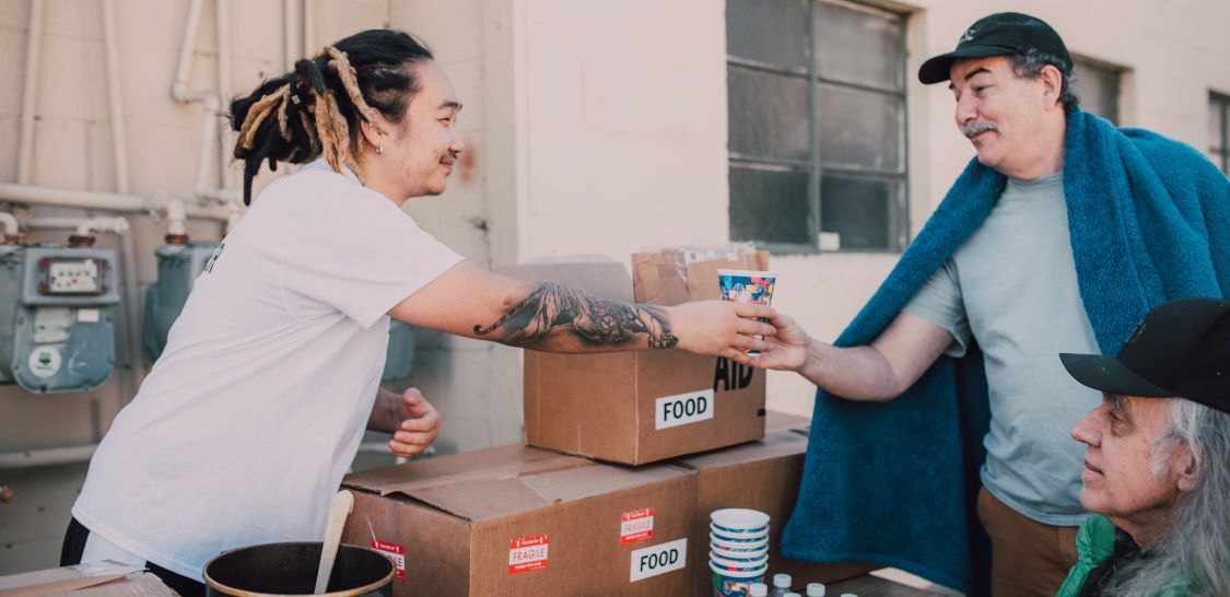 A man with dreadlocks hands a cup to a man wearing a blue baseball cap and a blanket draped around hi shoulders. There is a table full of boxes and small water bottles between them.