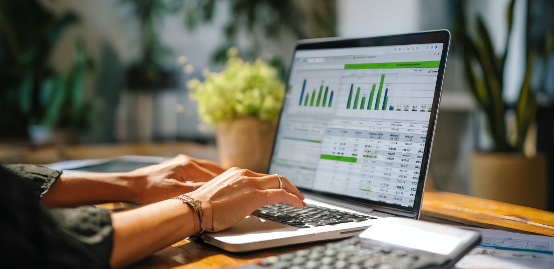 a person's hands are shown typing on a laptop with financial information on the screen. The laptop is sitting on a desk with a plant to the left.