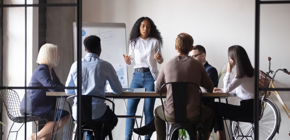 A woman wearing a white shirt, cardigan, and blue jeans stands in front of a table surrounded by multiple people with a large pad of paper on an easel behind her
