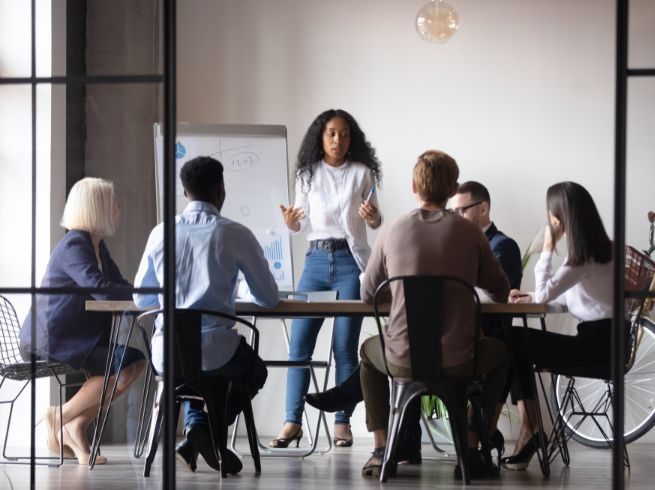 A woman wearing a white shirt, cardigan, and blue jeans stands in front of a table surrounded by multiple people with a large pad of paper on an easel behind her