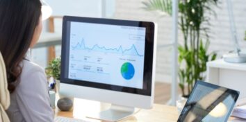 A woman sits at a desktop computer with data and a pie chart on the screen
