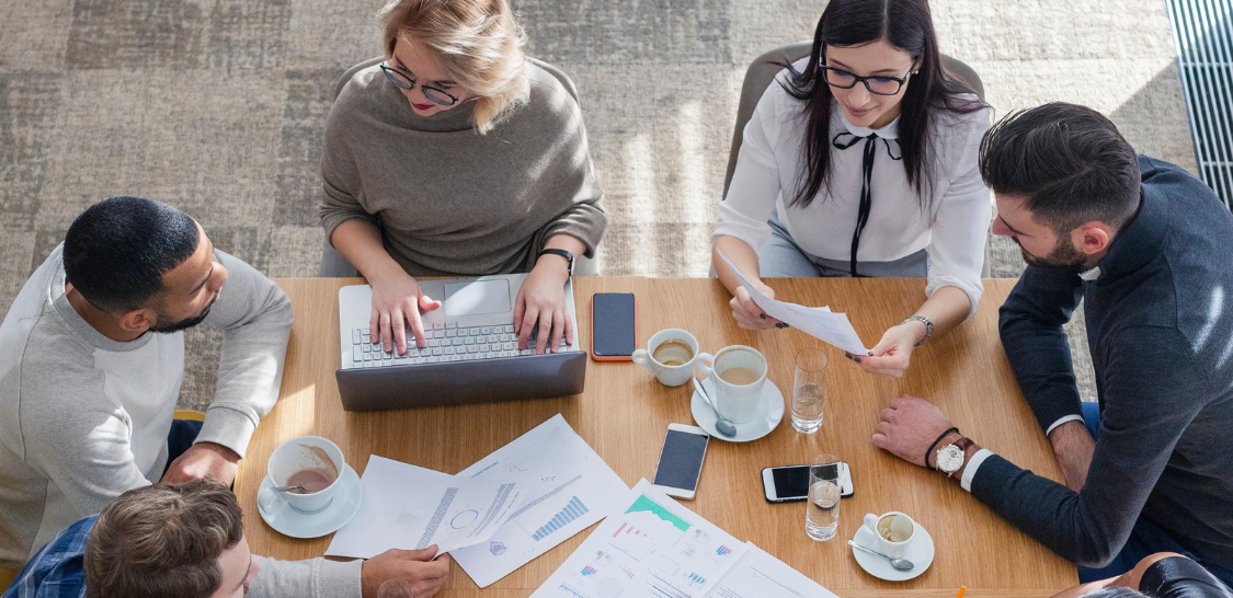 A group of diverse individual sit at a table discussing something, and there are papers and drinks and a laptop on the table. The image is photographed from overhead