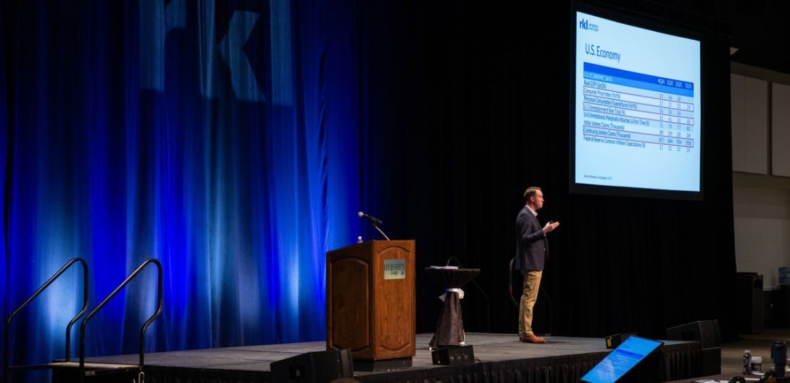 A man in a navy blue blazer and khaki pants stands on a stage giving a presentation with a podium on the left side of the image and a screen with a PowerPoint slide on the upper right of the image
