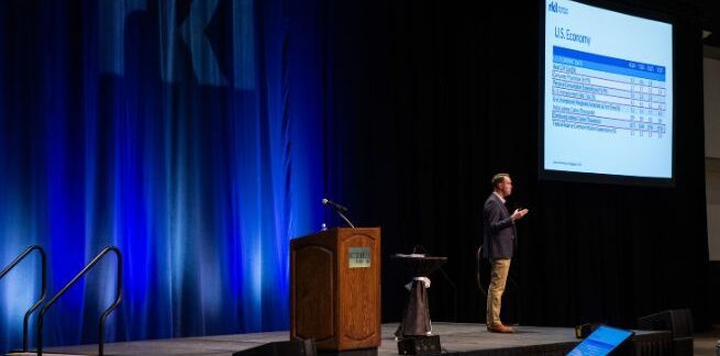 A man in a navy blue blazer and khaki pants stands on a stage giving a presentation with a podium on the left side of the image and a screen with a PowerPoint slide on the upper right of the image