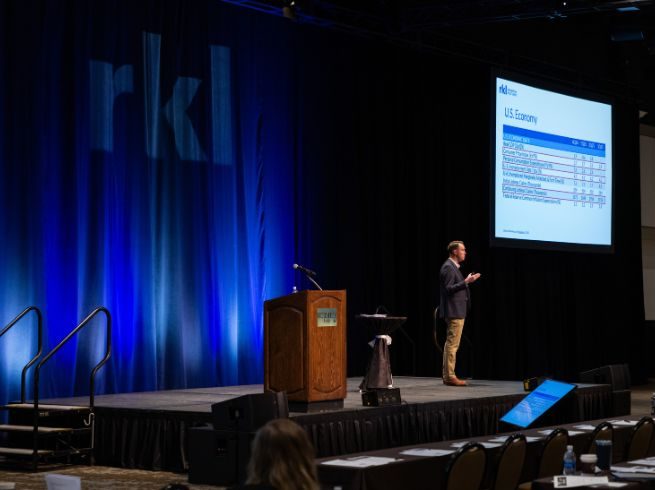 A man in a navy blue blazer and khaki pants stands on a stage giving a presentation with a podium on the left side of the image and a screen with a PowerPoint slide on the upper right of the image