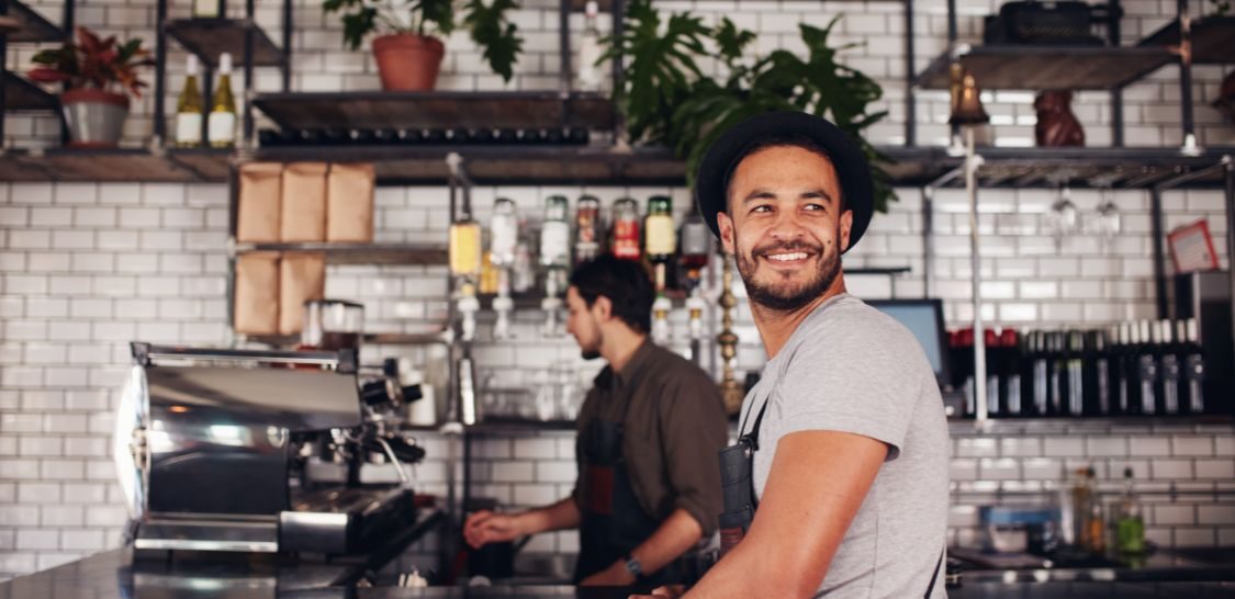 A man in a hat and apron leans against a counter at a coffee shop, and a man in a brown shirt wearing an apron makes coffee behind him