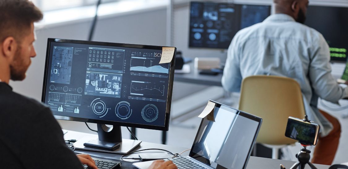 A man sits at a computer looking at the screen, which is filled with numbers and graphs and different things that would be on the screen of an information technology professional. There is another man in the background siting at a desk with a computer on it, looking at similar things on his screen.