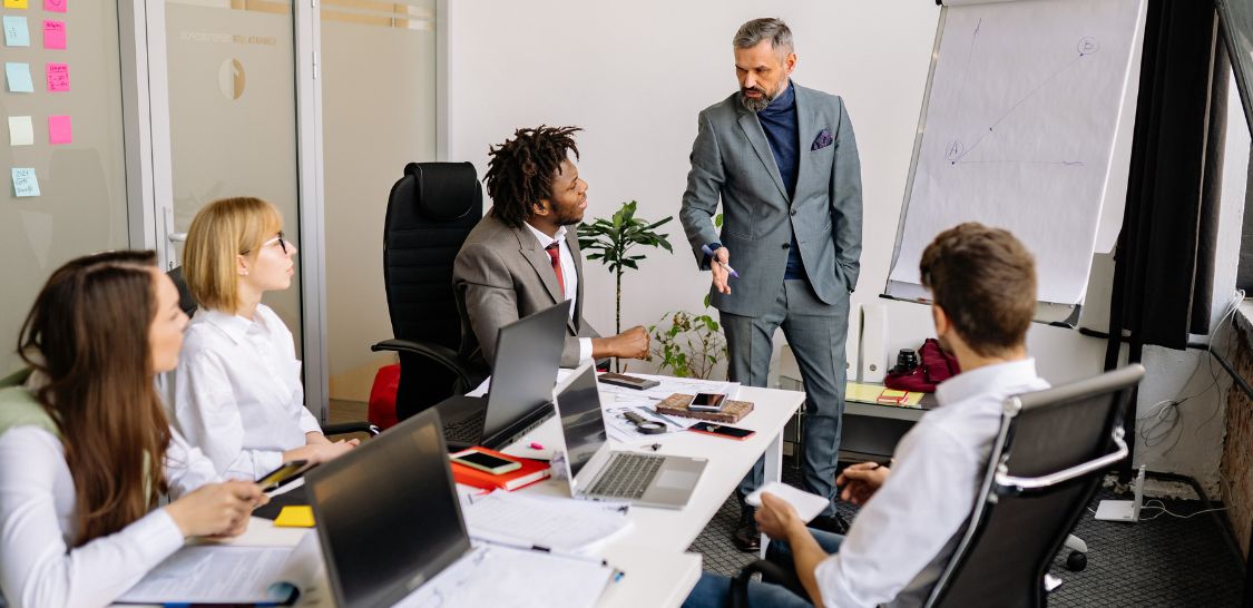 A group of diverse men and women sit around a table in a conference room where a man in a gray suit is giving a presentation and speaking to the people at the table. There is a large pad of paper on an easel at the front of the table with a graph on it.