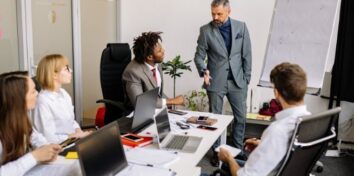 A group of diverse men and women sit around a table in a conference room where a man in a gray suit is giving a presentation and speaking to the people at the table. There is a large pad of paper on an easel at the front of the table with a graph on it.
