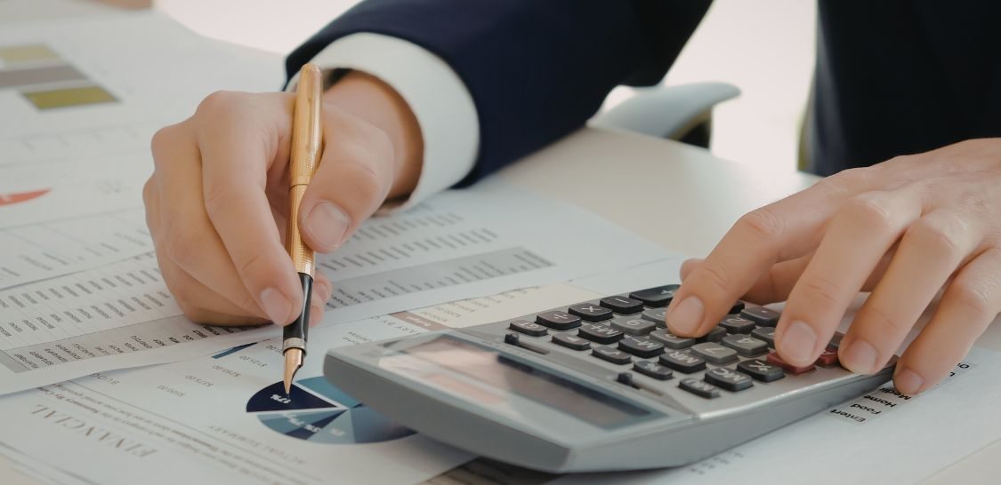 A person sits at a desk with a calculator and papers. Their hands are shown with their left hand typing on the calculator and their right hand holding a pen, getting ready to write on one of the papers.