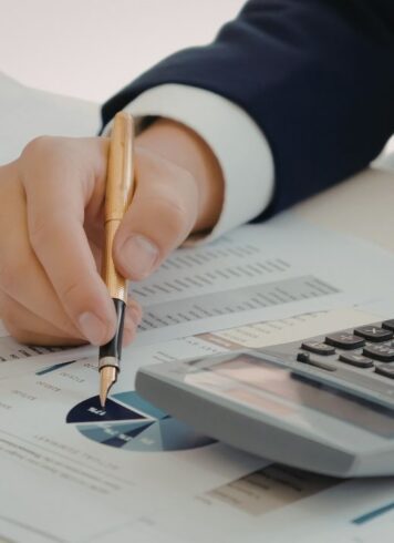 A person sits at a desk with a calculator and papers. Their hands are shown with their left hand typing on the calculator and their right hand holding a pen, getting ready to write on one of the papers.