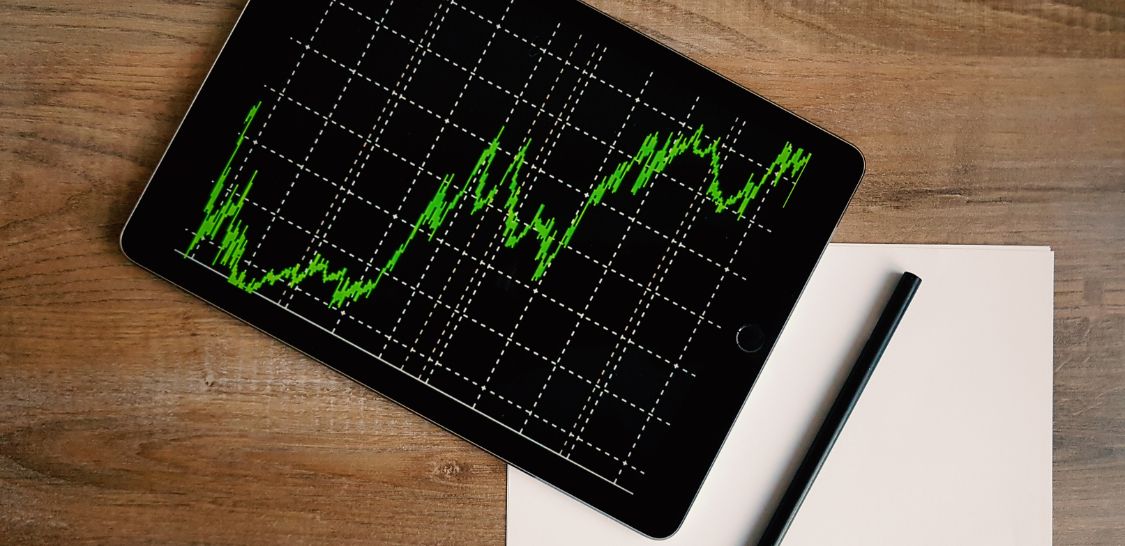 an electronic tablet with a black background and bright green line graph on the screen covers the corner of a piece of white paper with a pen sitting on it. All of this is on a wooden background, like a tabletop.
