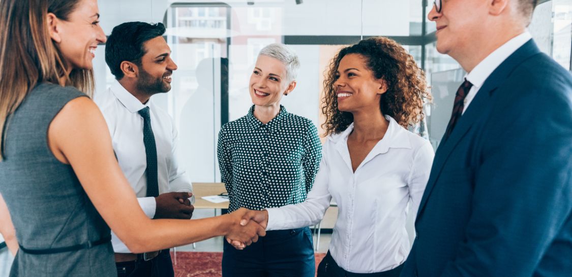 a diverse group of men and women wearing business attire stand in a circle. A woman in a white button down is shaking hands with a woman in a sage green sleeveless blouse