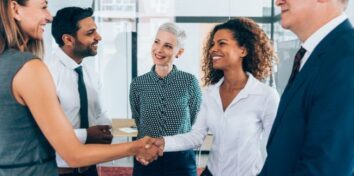 a diverse group of men and women wearing business attire stand in a circle. A woman in a white button down is shaking hands with a woman in a sage green sleeveless blouse