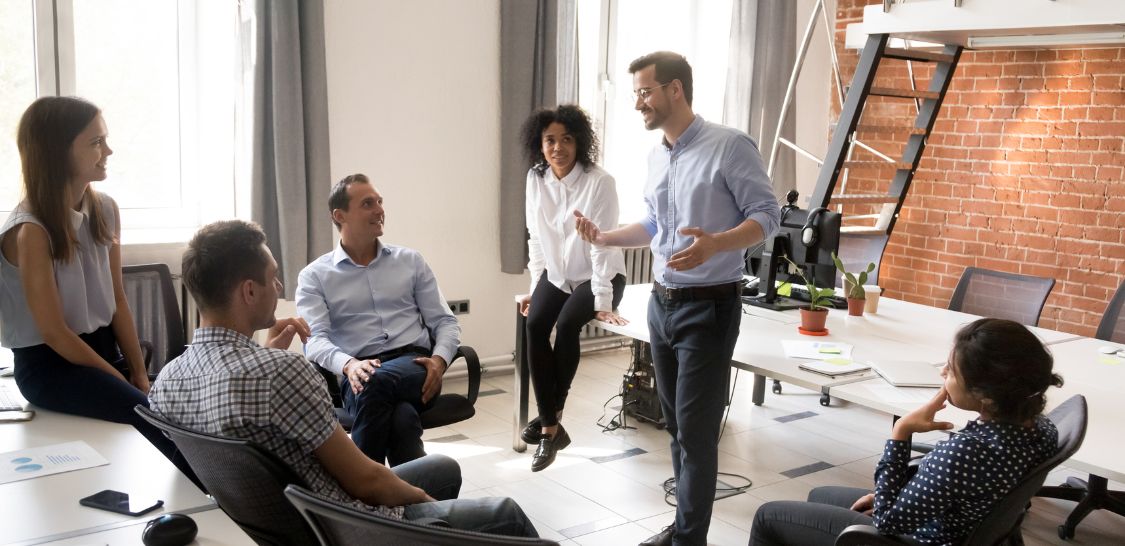 A diverse group of individuals in business clothing sit around a man who is talking.