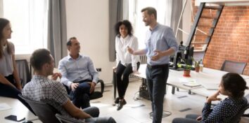 A diverse group of individuals in business clothing sit around a man who is talking.