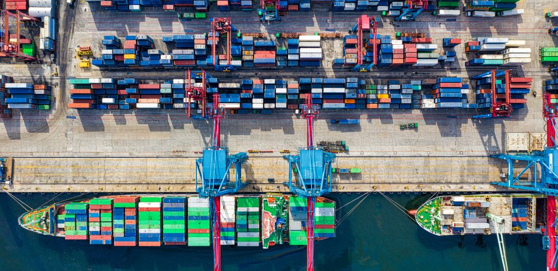 A photo from overhead of a port on the water with container ships at the top, two cranes in the center of the image loading containers onto a container ship