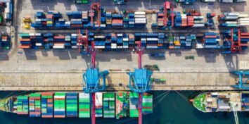 A photo from overhead of a port on the water with container ships at the top, two cranes in the center of the image loading containers onto a container ship