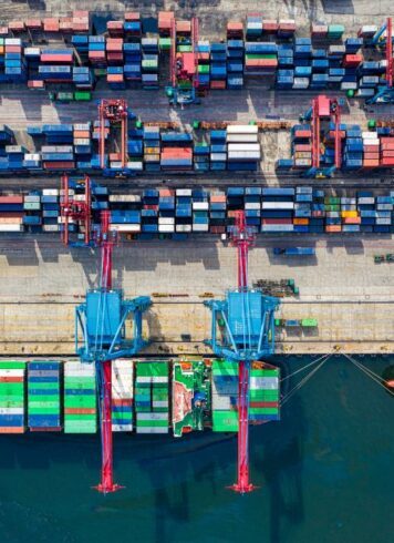 A photo from overhead of a port on the water with container ships at the top, two cranes in the center of the image loading containers onto a container ship