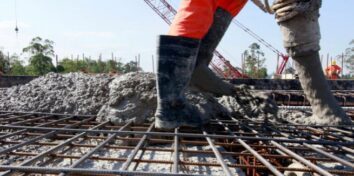 An image of a person from the knees down wearing rubber boots and orange construction uniform pants pouring cement over a rebar grid