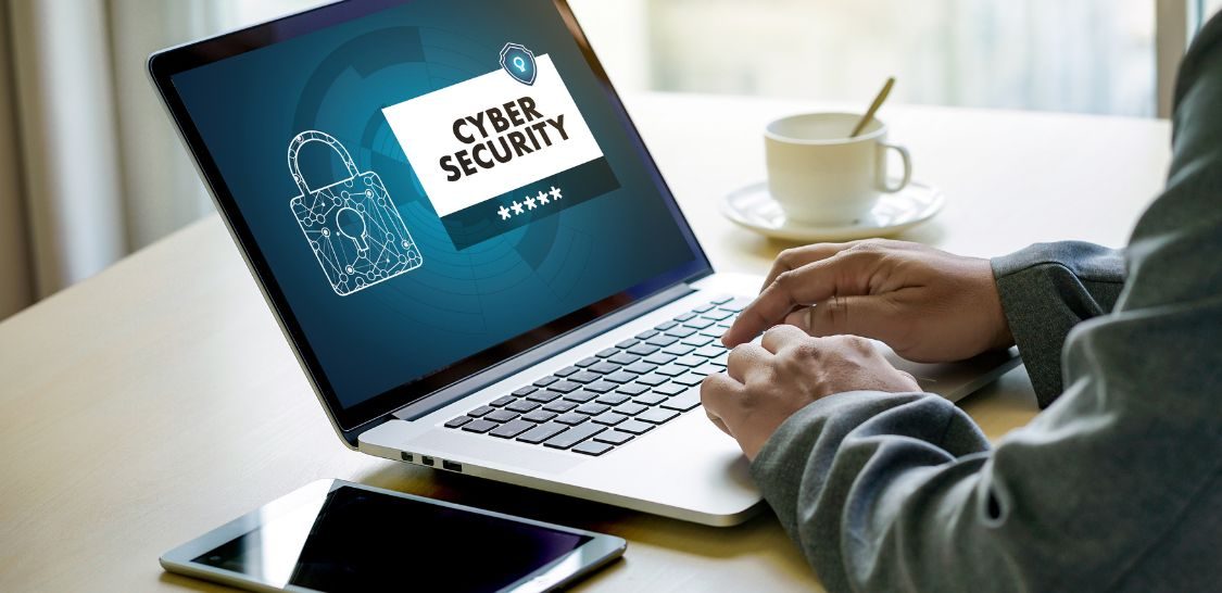 A laptop sits on a table and hands are typing on the keyboard. The screen has a lock on it and the words Cyber Security in a white box. A tablet sits on the table next to the laptop.
