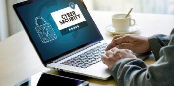 A laptop sits on a table and hands are typing on the keyboard. The screen has a lock on it and the words Cyber Security in a white box. A tablet sits on the table next to the laptop.