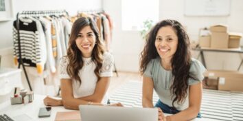 Two women sit at a desk with a laptop in front of them and a rack of clothing behind them to the left