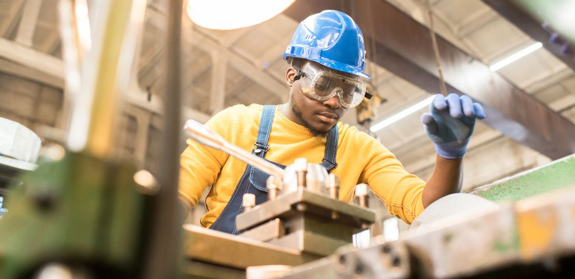 A man in a yellow shirt and blue hardhat and goggles stands over a machine, working