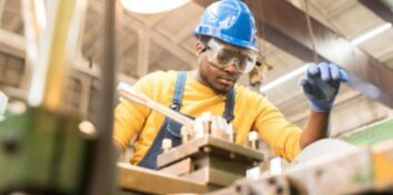 A man in a yellow shirt and blue hardhat and goggles stands over a machine, working