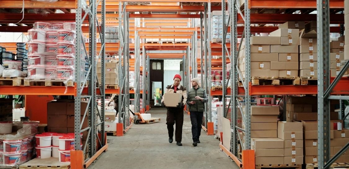 A man carrying a box next to a man carrying a tablit walk through a warehouse with shelves of boxes on either side of them