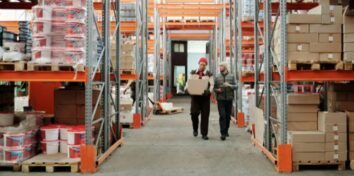 A man carrying a box next to a man carrying a tablit walk through a warehouse with shelves of boxes on either side of them