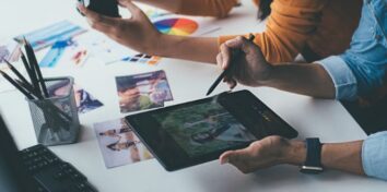 A close up photo of two sets of hands over a table. One person is holding a camera while the other has a stylus over an electronic tablet, editing a photo