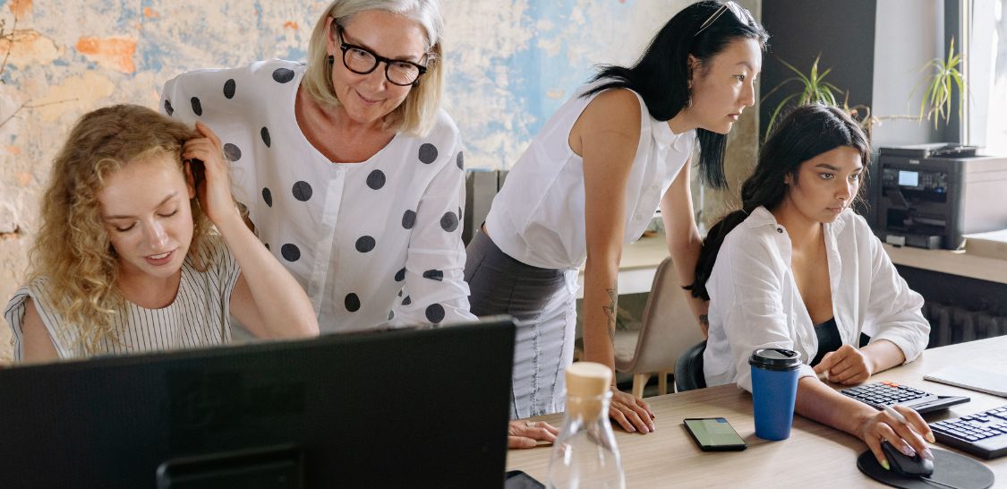 Four women work at a desk with two sitting working on a laptop while two others stand behind them appearing to help them with their work.