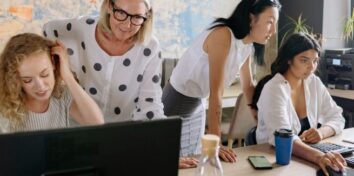 Four women work at a desk with two sitting working on a laptop while two others stand behind them appearing to help them with their work.