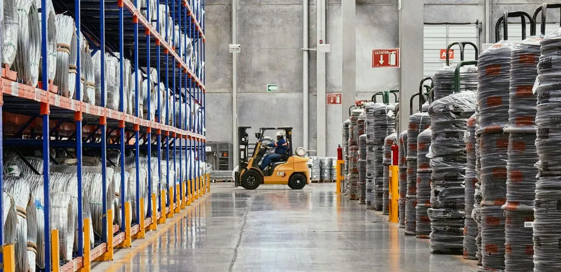 A man drives a forklift at the back of a warehouse and is being photographed from afar between shelves of large wire spools
