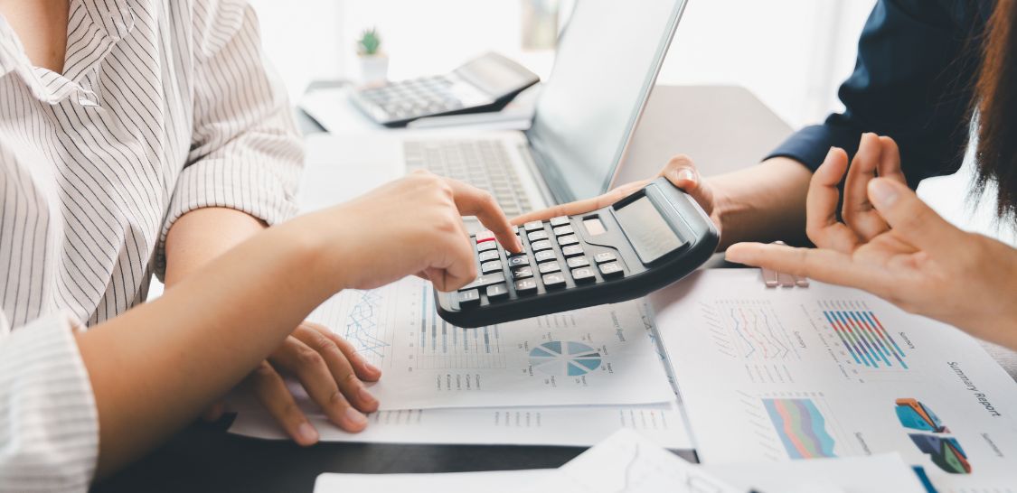 A person sits at a desk and types on a calculator that another person is holding. There are papers with graphs and charts on the table as well as a laptop.