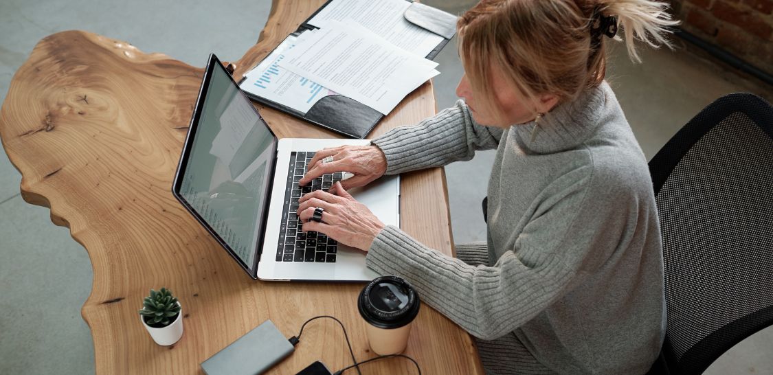 A woman sits at a table typing on a laptop with papers on her right and a coffee cup and cell phone on her left