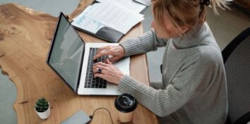 A woman sits at a table typing on a laptop with papers on her right and a coffee cup and cell phone on her left