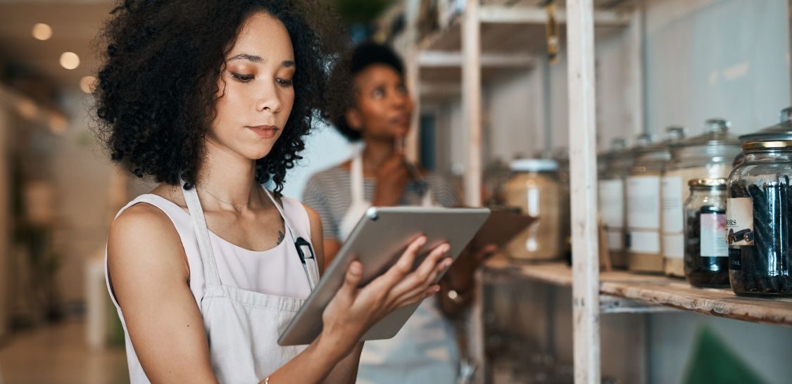 A woman holding a tablet stands in front of a shelf of items with a woman in the background looking at the shelf