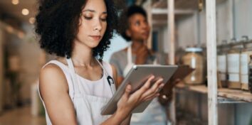 A woman holding a tablet stands in front of a shelf of items with a woman in the background looking at the shelf