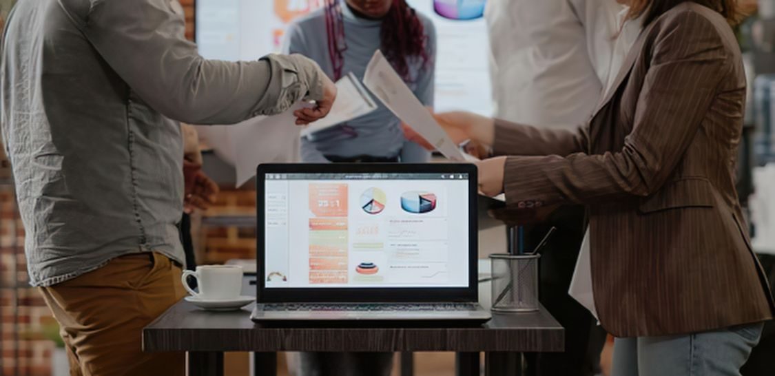A laptop sits on a desk facing the camera with charts and graphs on the screen. Two people stand in the background and they are exchanging information on paper.