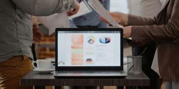 A laptop sits on a desk facing the camera with charts and graphs on the screen. Two people stand in the background and they are exchanging information on paper.